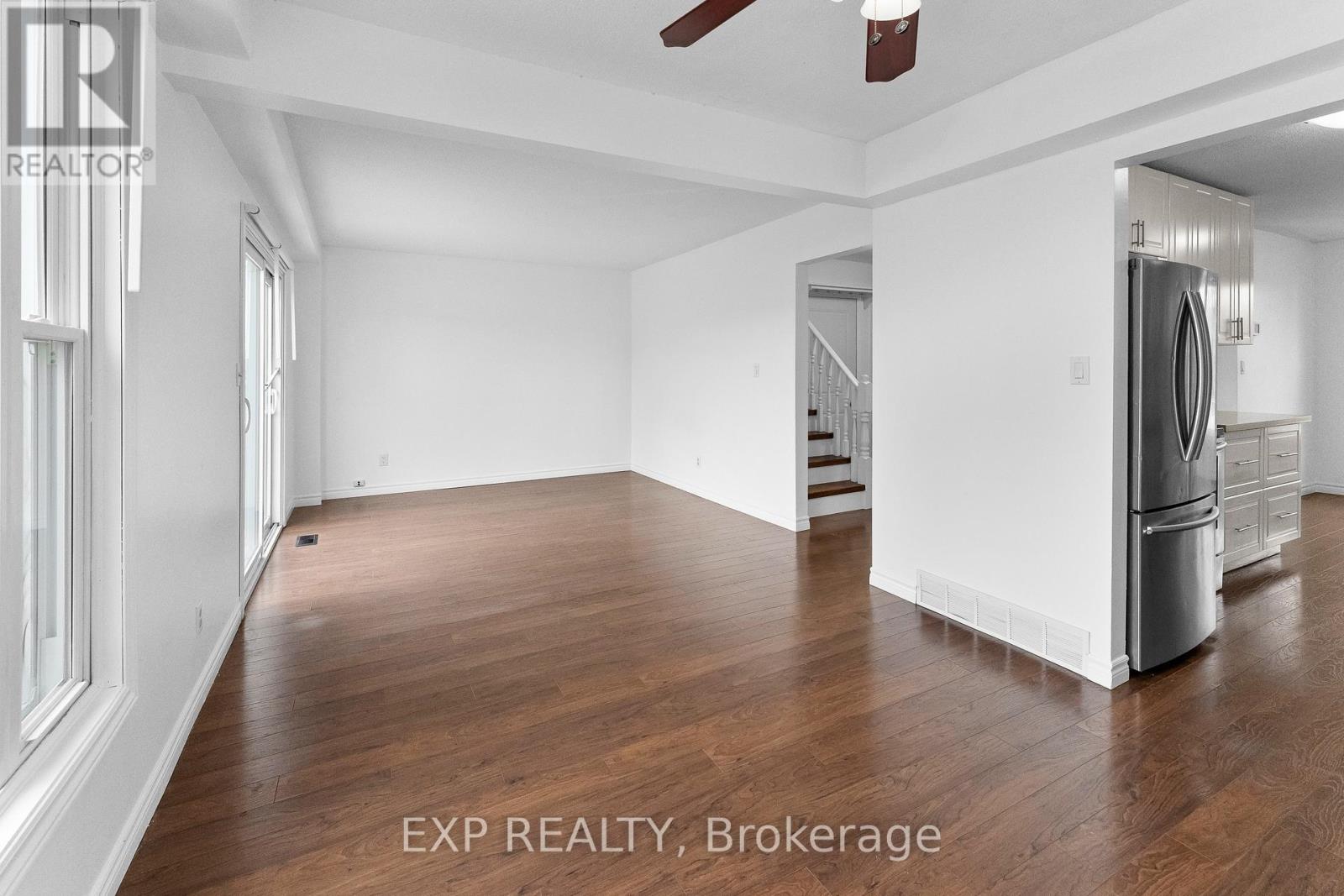 A spacious room with wooden flooring and white walls, with a staircase in the background.