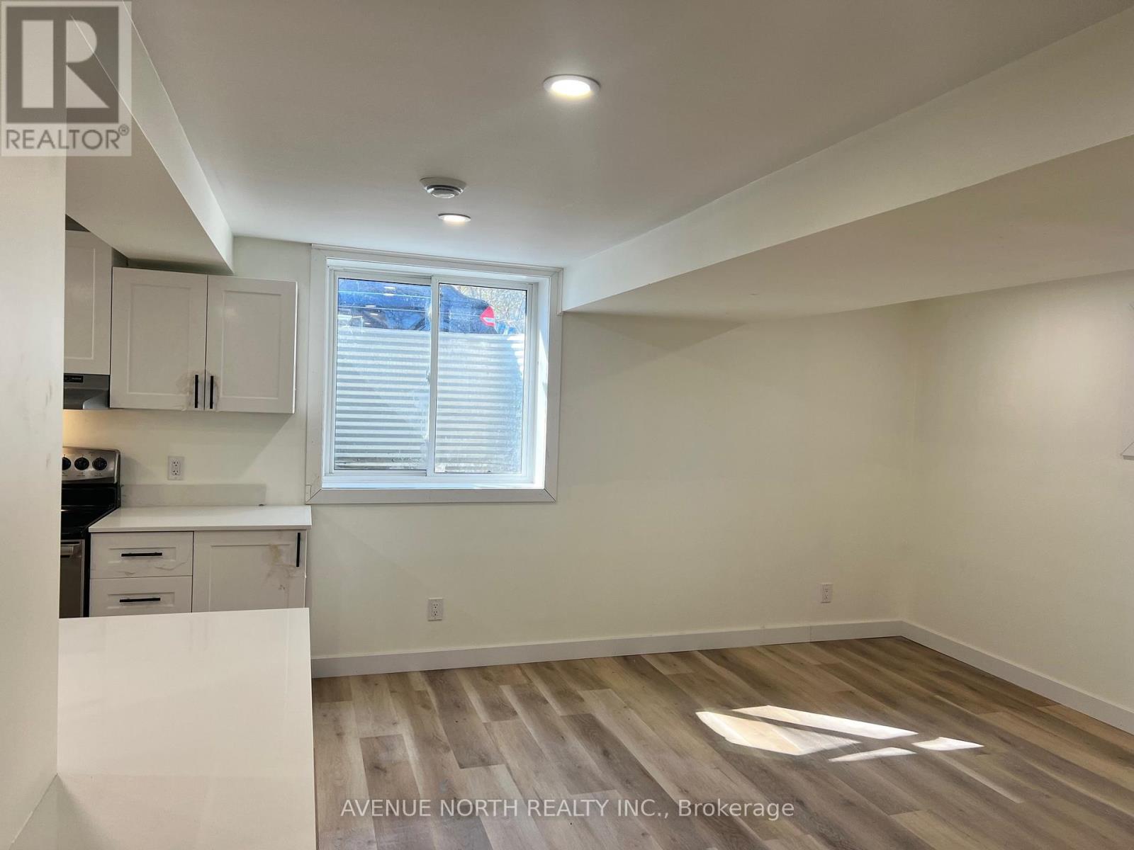 A real estate listing photo of a white kitchen with wood flooring and a window.