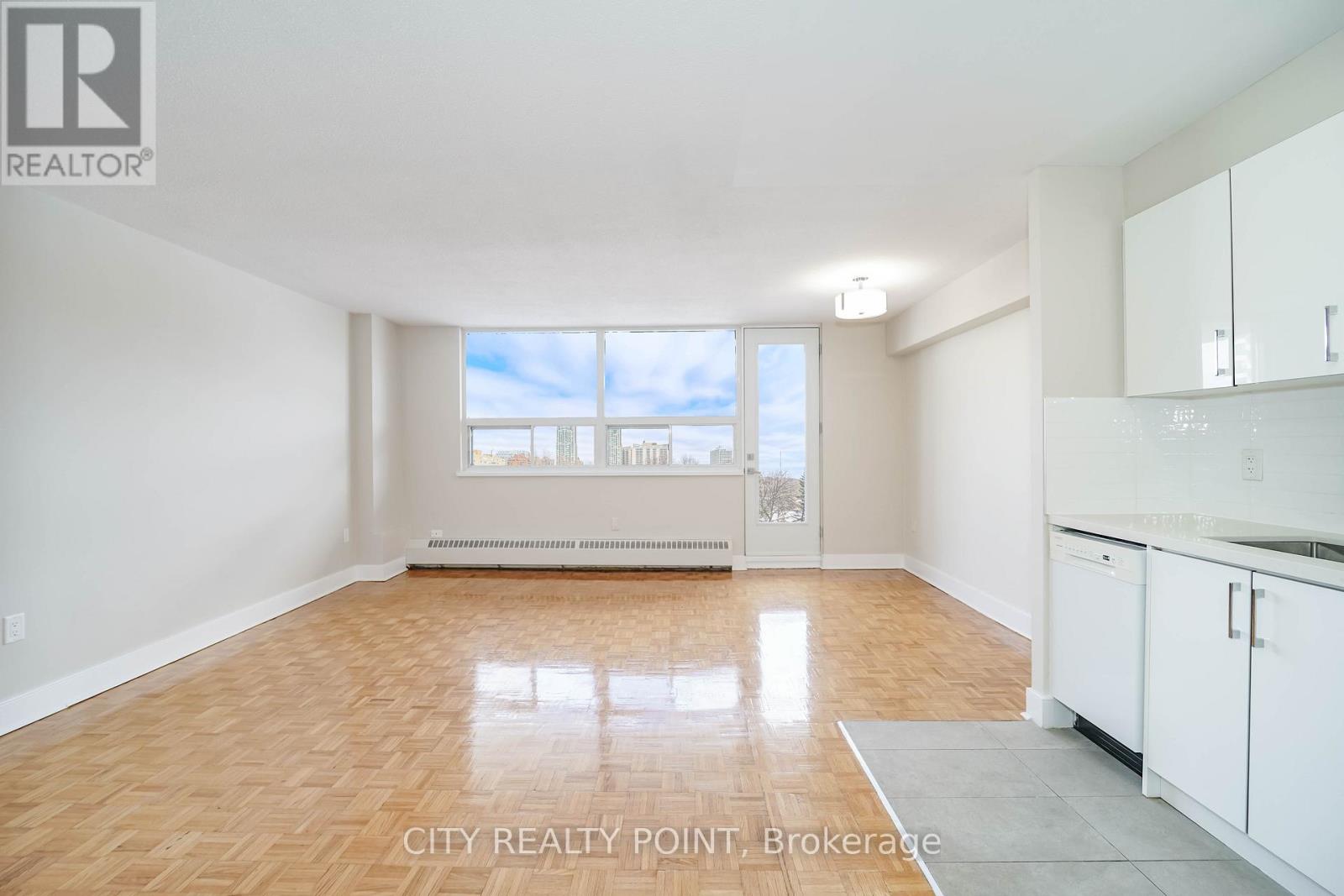 A spacious room with wooden flooring and white walls, with a view of a cityscape through the windows.