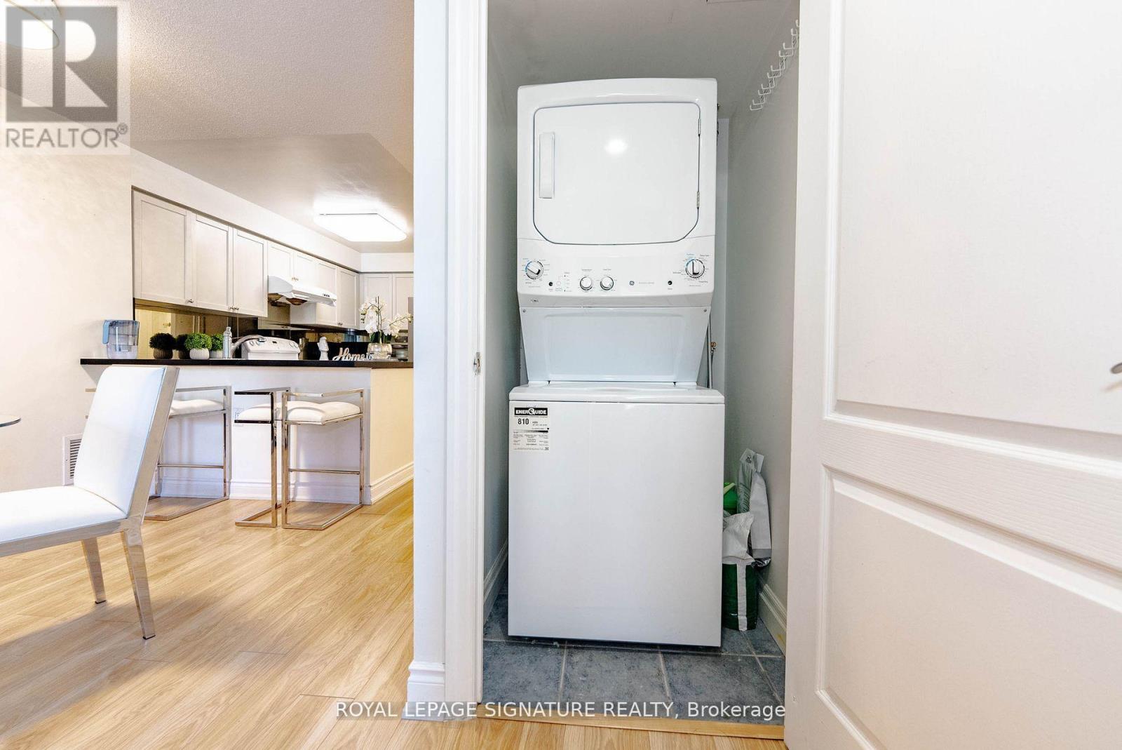 A white washing machine is in a small room with a white chair and a small kitchen.