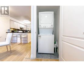 A white washing machine is in a small room with a white chair and a small kitchen.