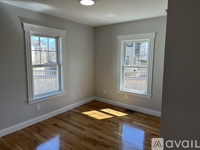A room with wooden floors and two windows, one of which has a view of a fence and buildings outside.