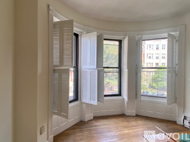 A room with white walls and wooden flooring, featuring a bay window with white shutters.