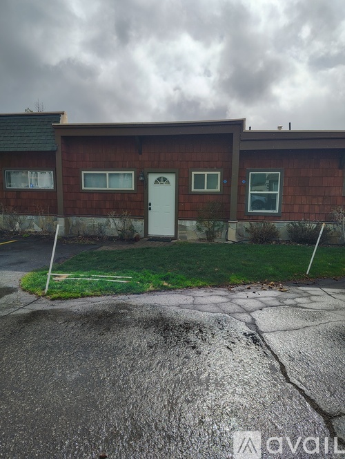 A house with a red brick exterior and a white door is surrounded by a grassy area and a cracked driveway.