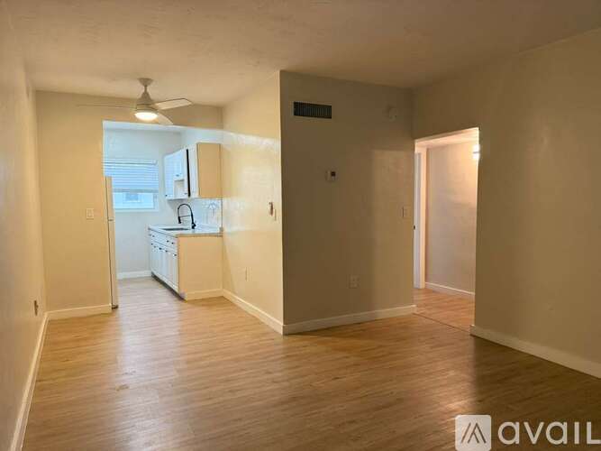 A room with wooden floors and a kitchen area in the background.