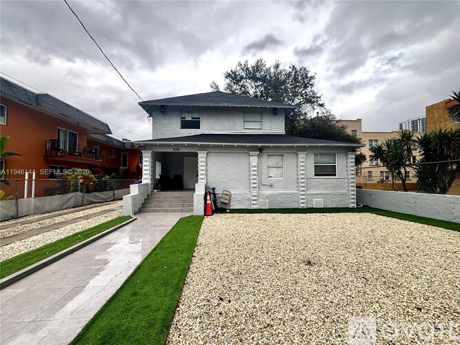 A house with a grey roof and white walls is surrounded by a gravel driveway and a grassy area.