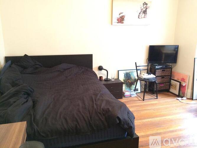 A bed with a black comforter is in the foreground of a room with a television and a chair.