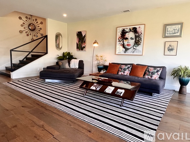 A living room with a black and white striped rug, a black couch with orange pillows, and a staircase with a metal railing.