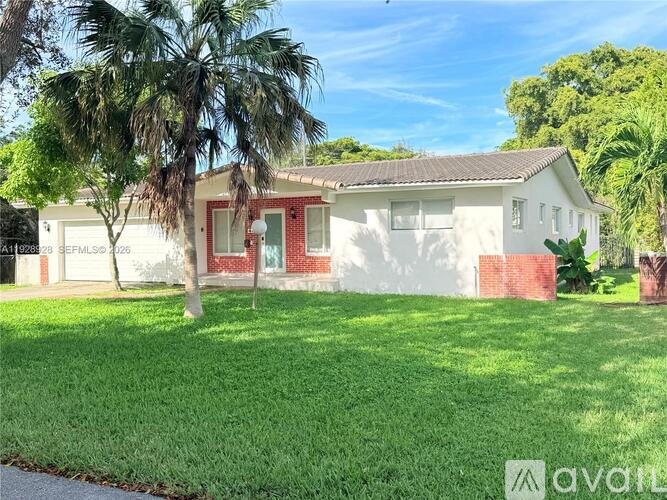 A house with a red door and a white wall is surrounded by green grass and trees.