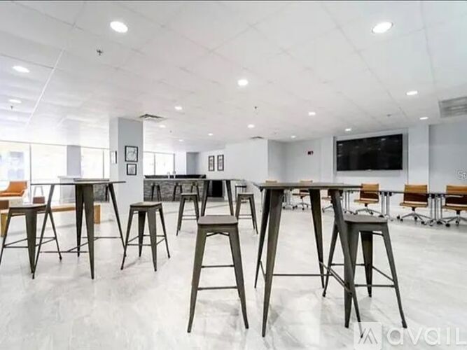A modern office space with a white floor and white walls, featuring a long table with chairs and a TV mounted on the wall.
