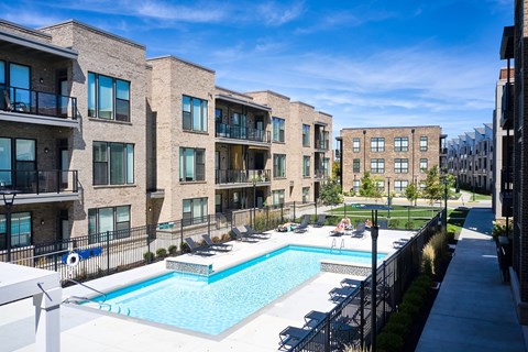 A swimming pool is surrounded by lounge chairs in front of apartment buildings.