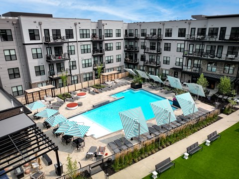 A large swimming pool surrounded by lounge chairs and umbrellas in front of apartment buildings.