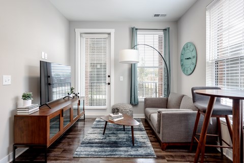 A living room with a grey couch, a wooden cabinet, a coffee table, a lamp, a clock, and a window with blinds.