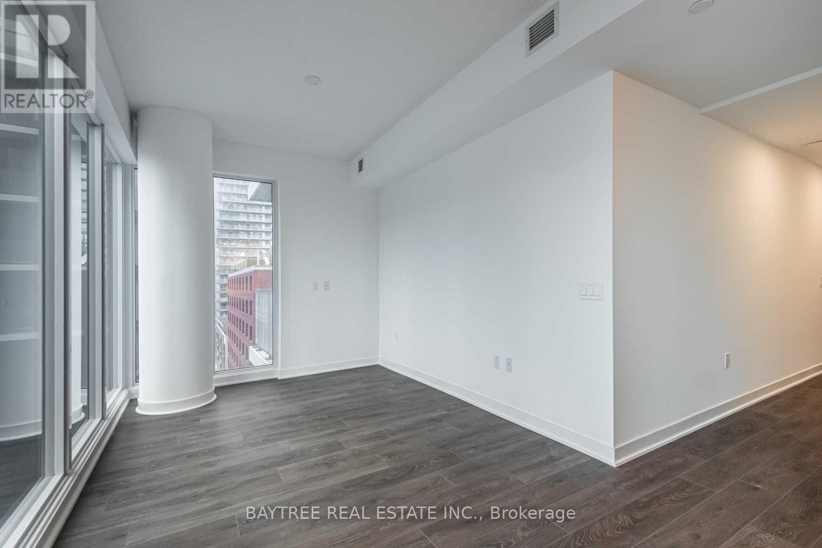 A spacious room with wooden flooring and white walls, with a window that offers a view of a building outside.