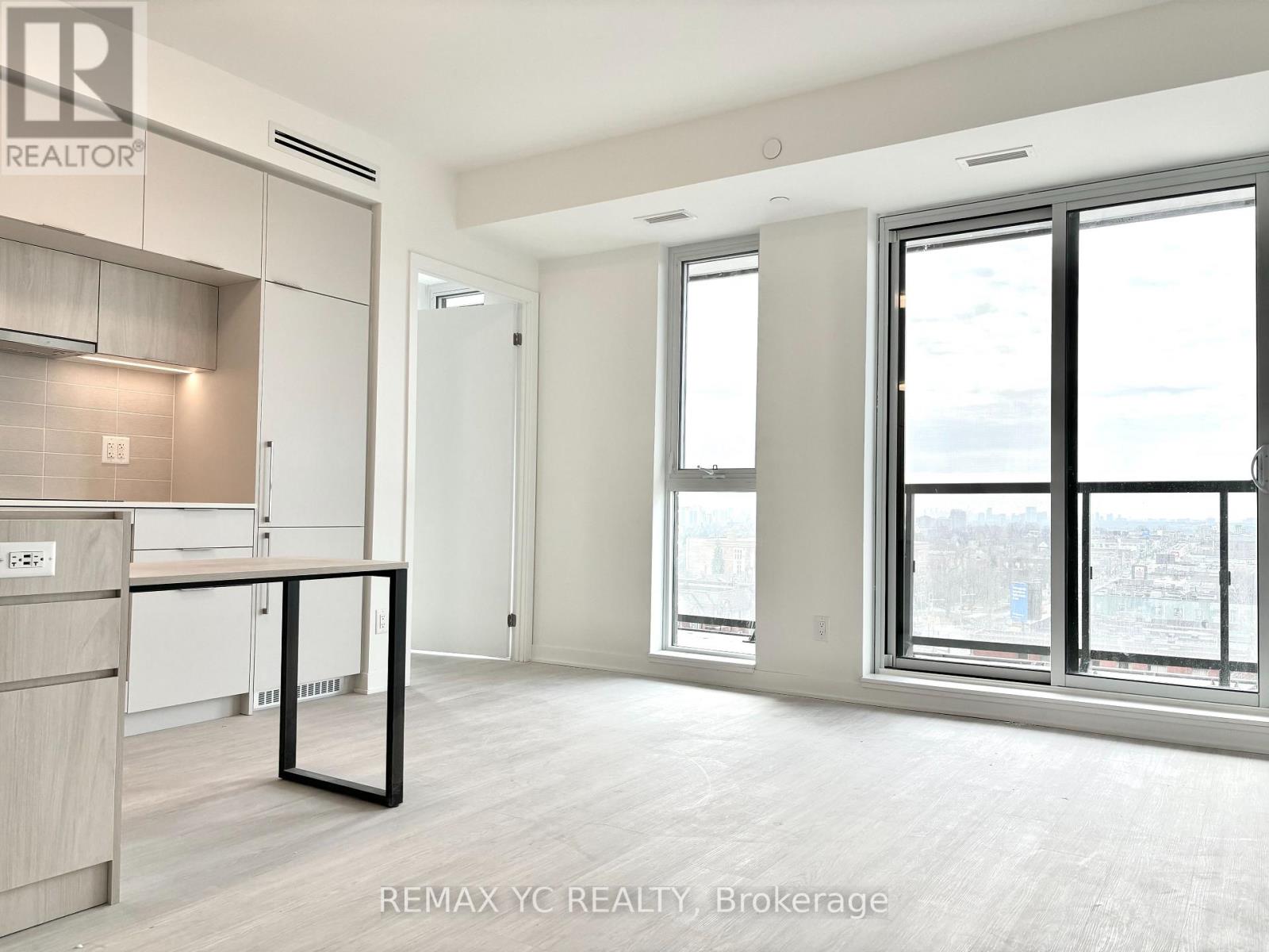 A spacious, empty room with a kitchen area to the left and a large window with a view of the city.