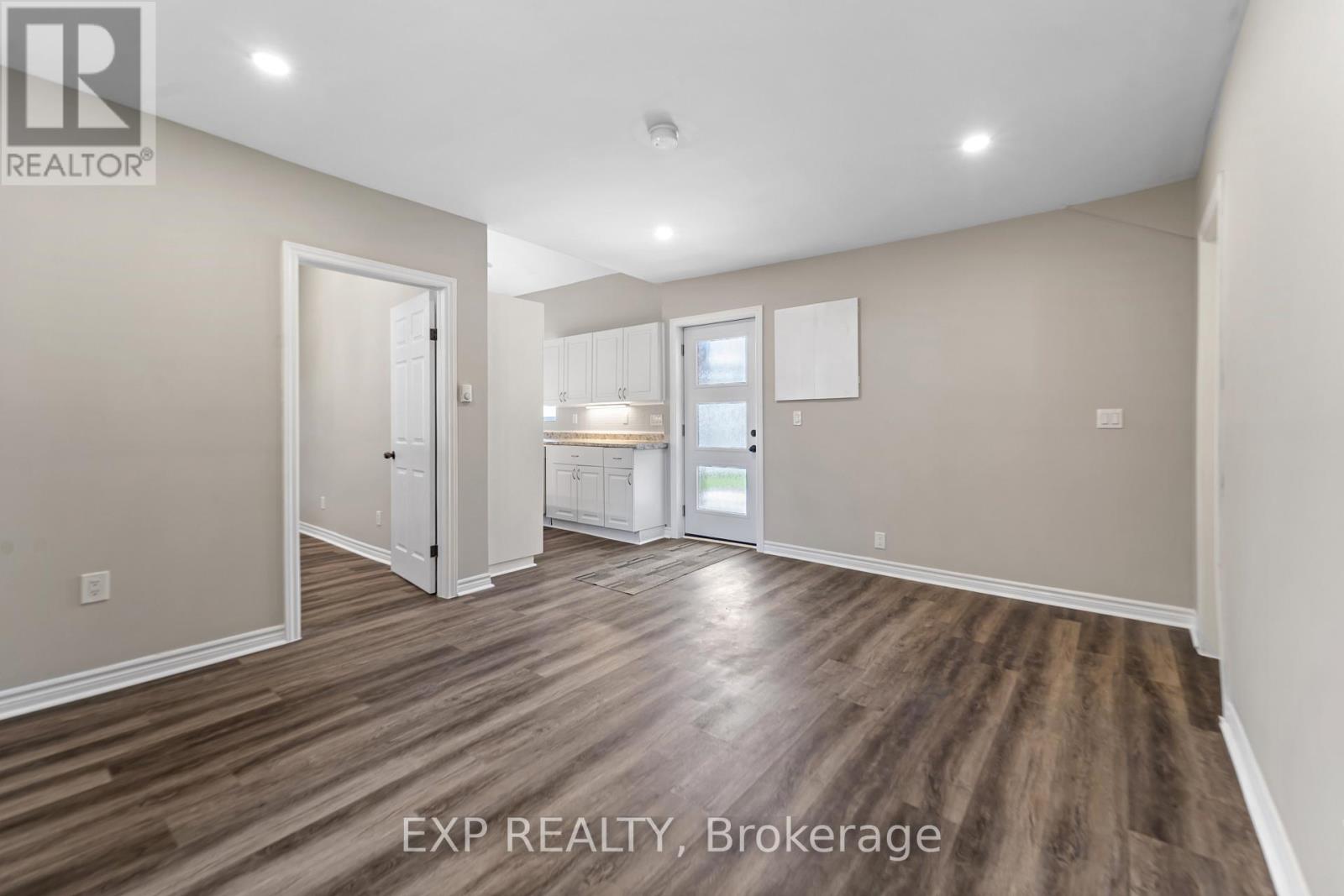 A room with wood flooring and white walls, with a door on the left and a kitchenette on the right.