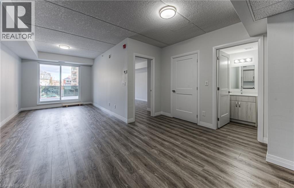 A spacious room with wooden flooring and white walls, featuring a large window, a door, and a small bathroom with a mirror.