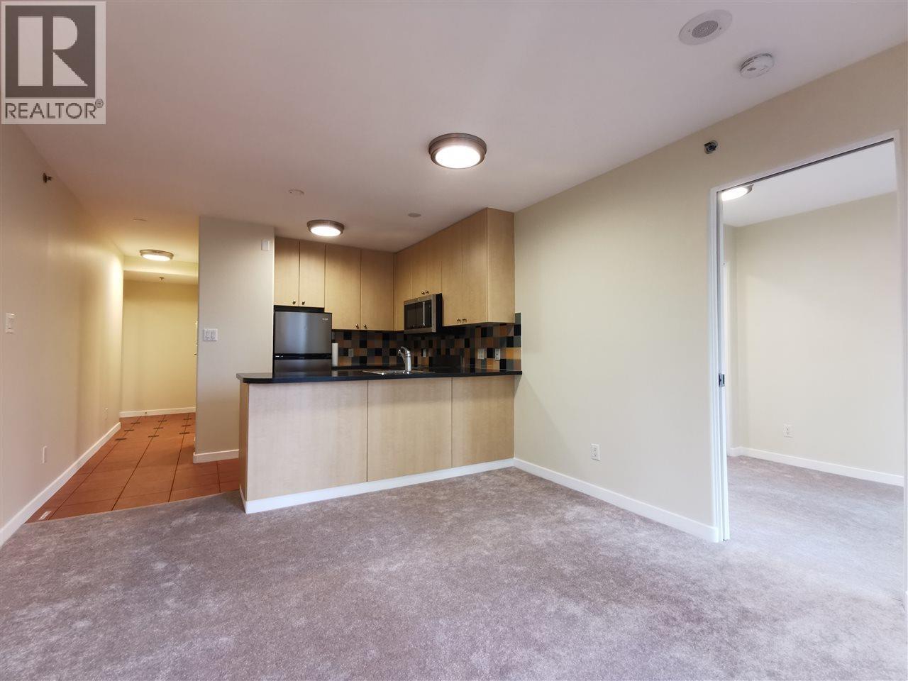 A well-lit, empty kitchen and living room with a grey carpet.