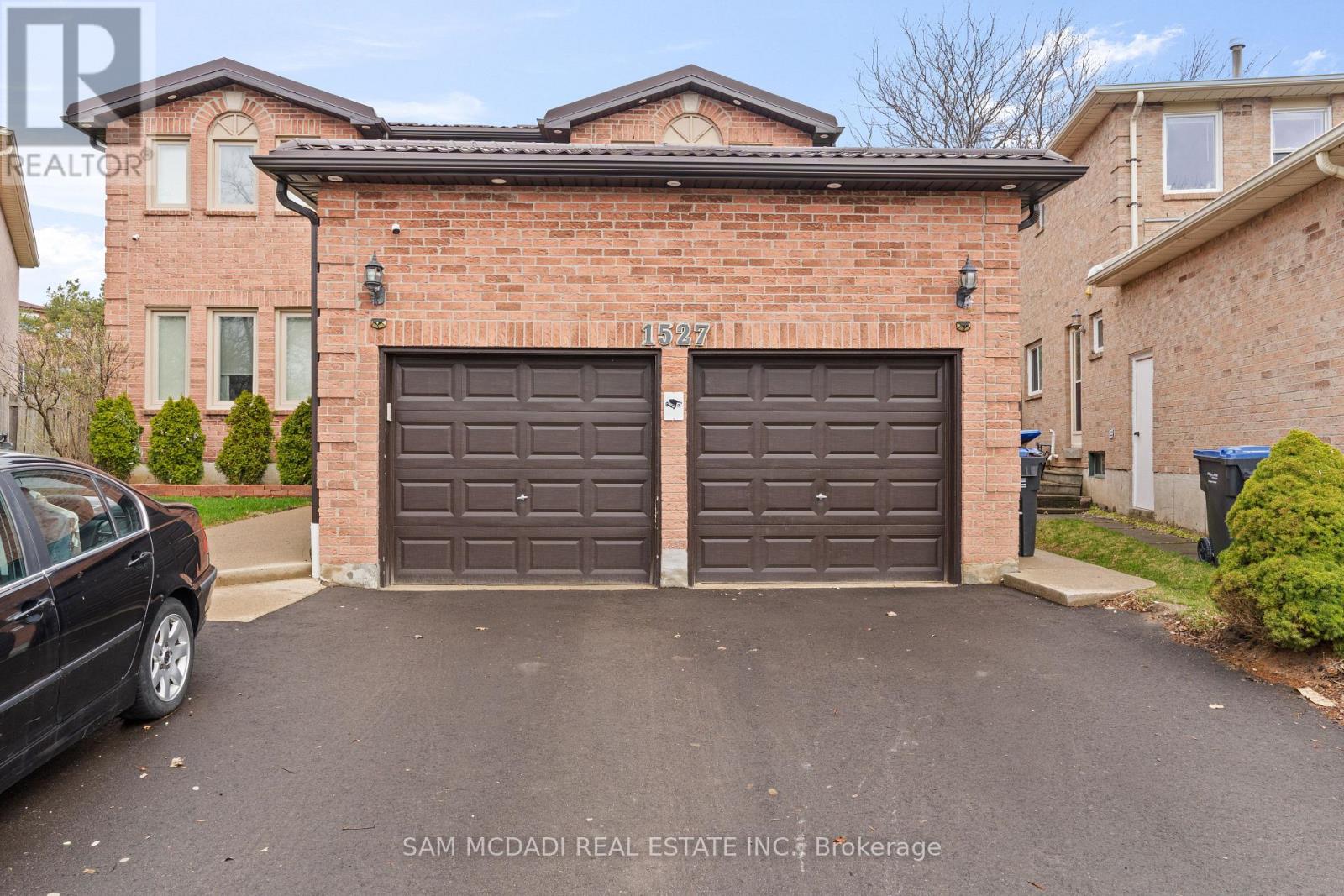 A black car is parked in front of a brick house with a brown garage door.
