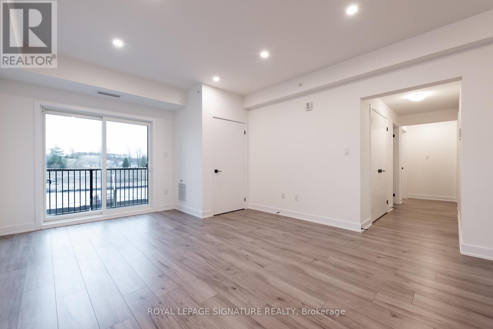 A spacious room with wooden flooring and white walls, with a balcony visible through the window.