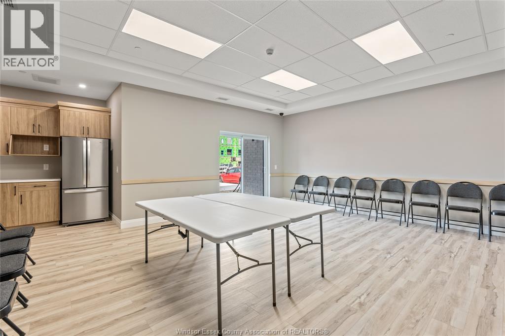A conference room with a white table and chairs, and a kitchen area with a refrigerator and cabinets in the background.