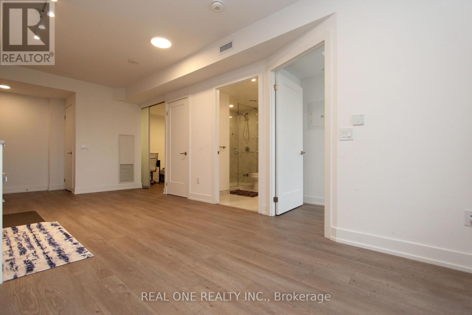 A spacious room with wooden flooring and white walls, featuring a bathroom with a glass shower door.