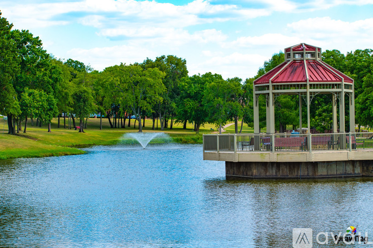 A gazebo sits on a pier over a lake with a fountain in the foreground.