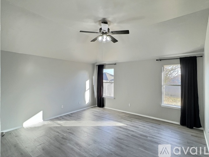 A spacious room with a ceiling fan and light, hardwood floors, and a window with black curtains.