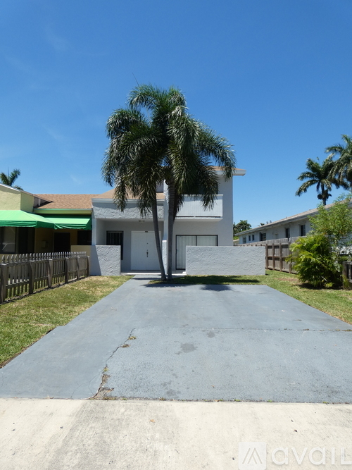 A white building with a green awning is surrounded by a fence and a tall palm tree.