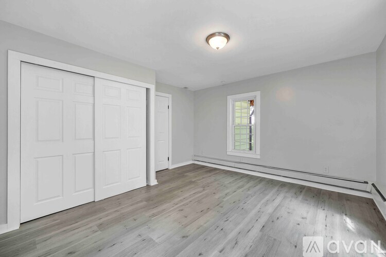 A spacious room with a white door, a window, and wooden flooring.