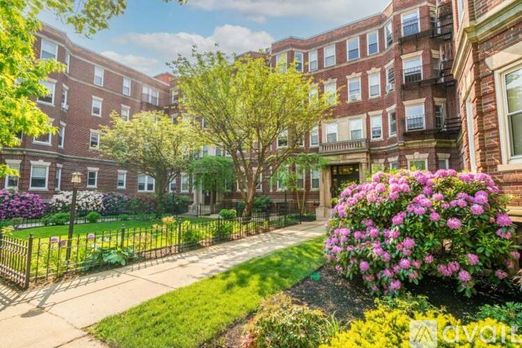 A sunny day at a brick apartment complex with a tree and flower bed in the foreground.