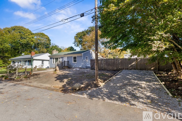 A gravel driveway leads to a small house with a fence and trees in the background.