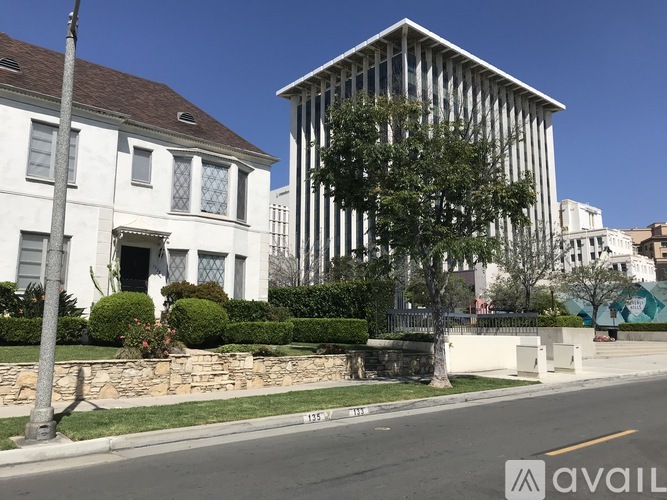 A white building with a brown roof is in front of a tall white building with a tree in front of it.