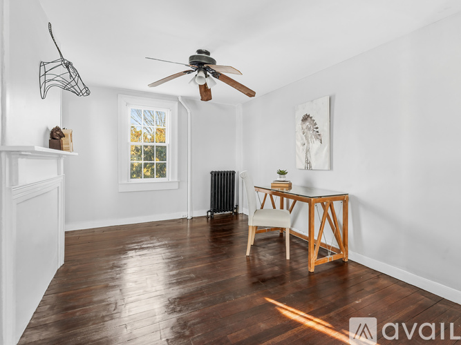 A room with a wooden floor, a ceiling fan, a table with a chair, and a window with a view of trees.