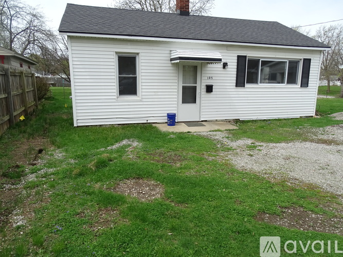 A small white house with a black roof and a blue trash can in front of it.