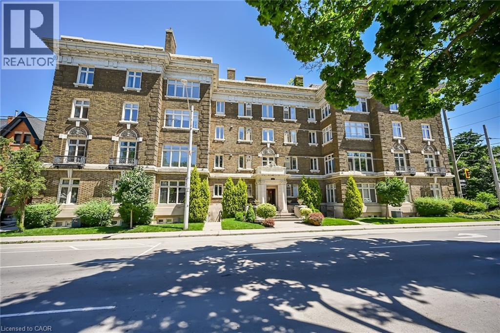 A large, multi-story building with a stone facade and multiple windows is surrounded by greenery and a clear sky.