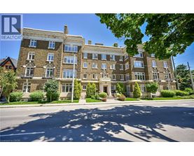 A large, multi-story building with a stone facade and multiple windows is surrounded by greenery and a clear sky.