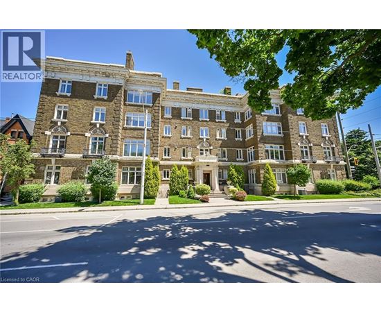 A large, multi-story building with a stone facade and multiple windows is surrounded by greenery and a clear sky.