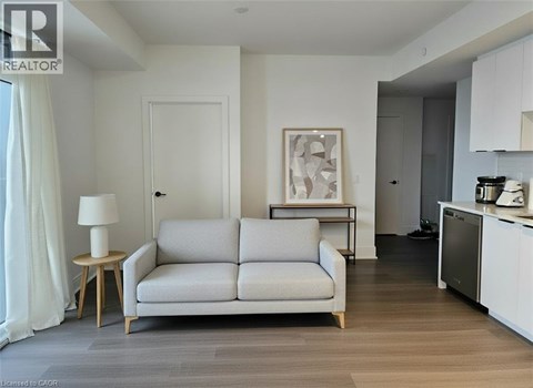 A living room with a grey couch, a side table with a lamp, and a kitchen area with a fridge and stove.