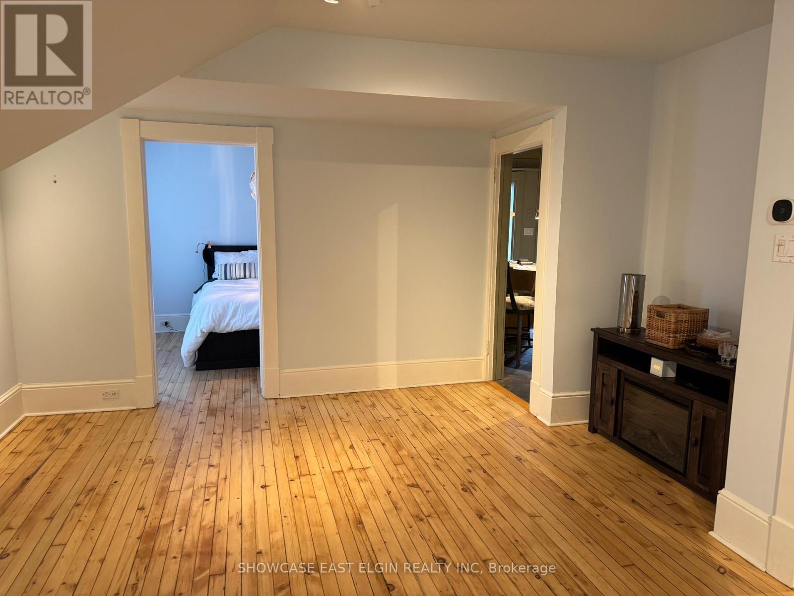 A room with wooden flooring and white walls, with a bed visible through an open door.
