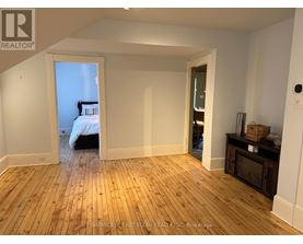 A room with wooden flooring and white walls, with a bed visible through an open door.