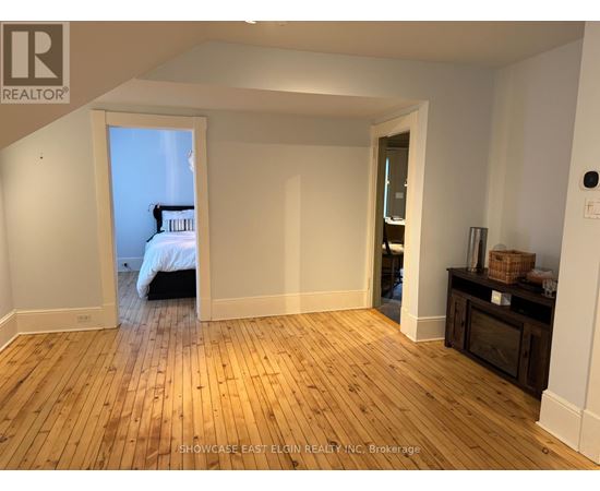 A room with wooden flooring and white walls, with a bed visible through an open door.
