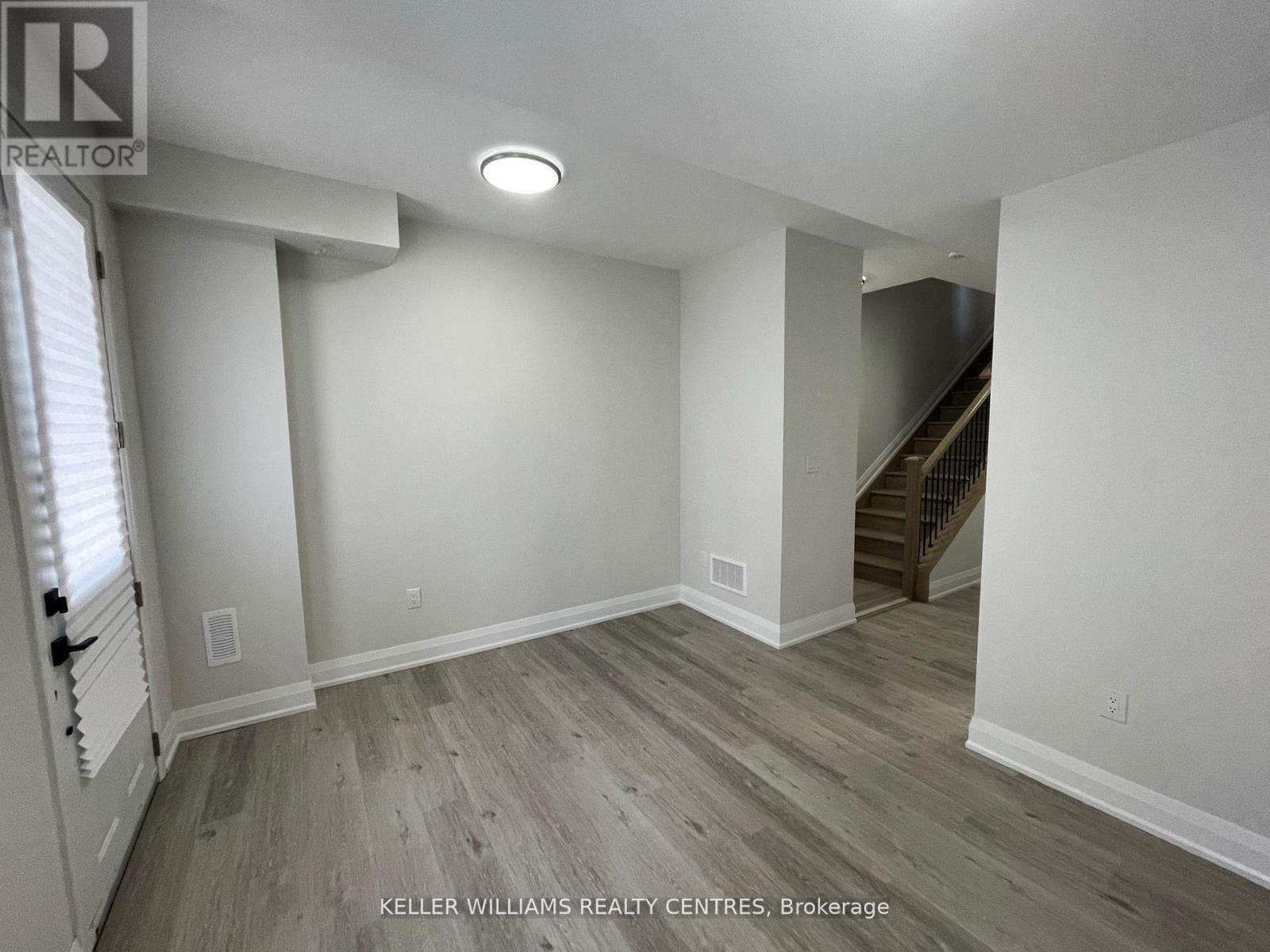 A spacious room with wood flooring and a staircase in the background.