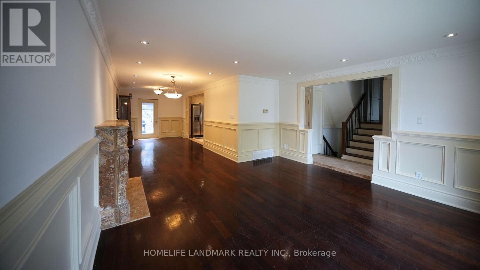 A spacious room with wooden floors and white walls, featuring a staircase and a chandelier.