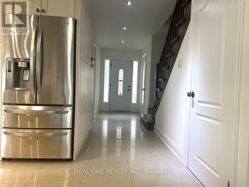 A stainless steel refrigerator is in the foreground of a hallway with white walls and a staircase.