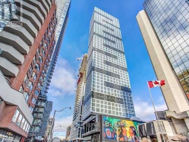 A tall glass building stands in the middle of a city street, with a Canadian flag flying in front of it.