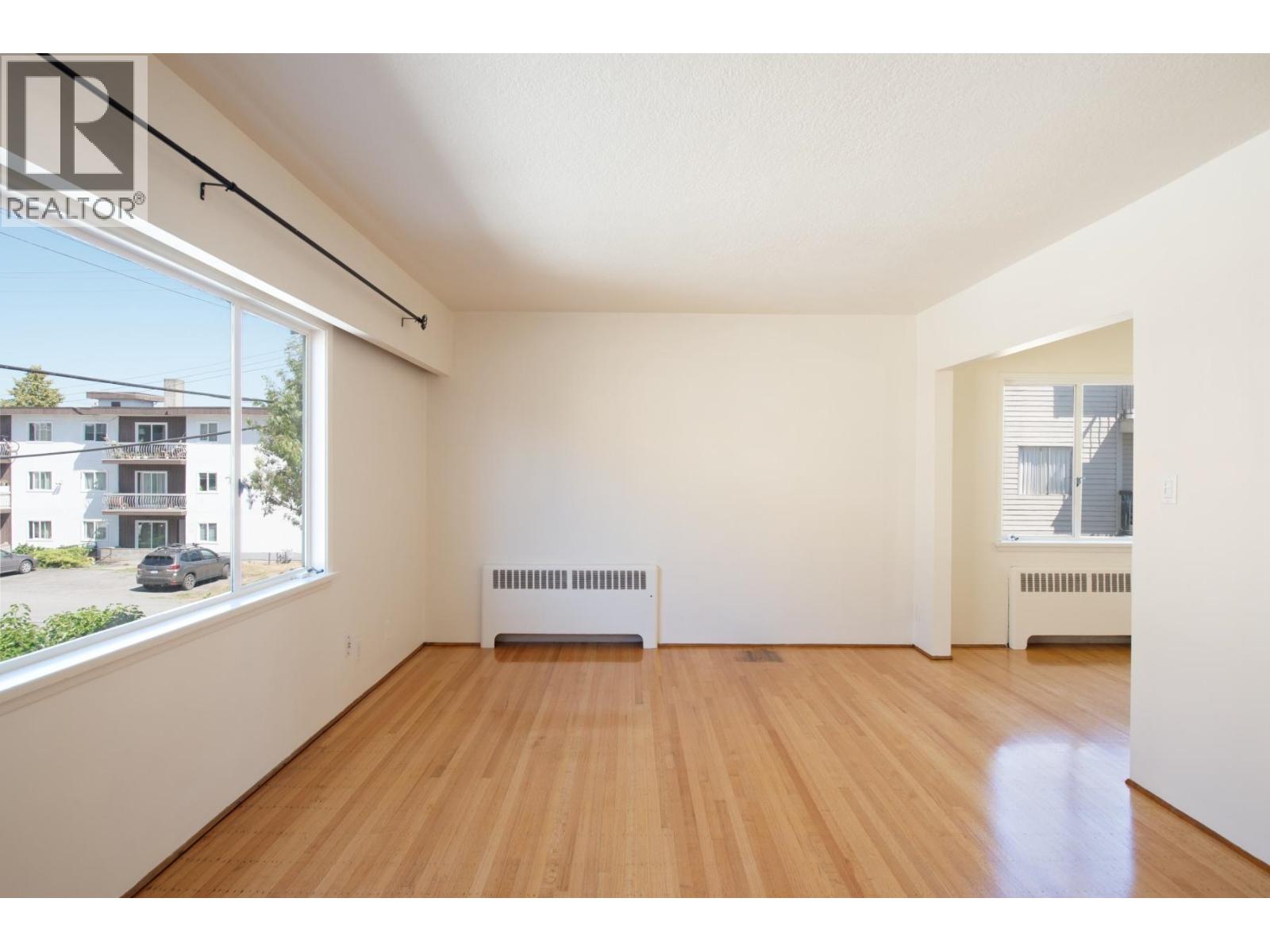A room with wooden floors and white walls, with a window showing a view of a street with cars and buildings.