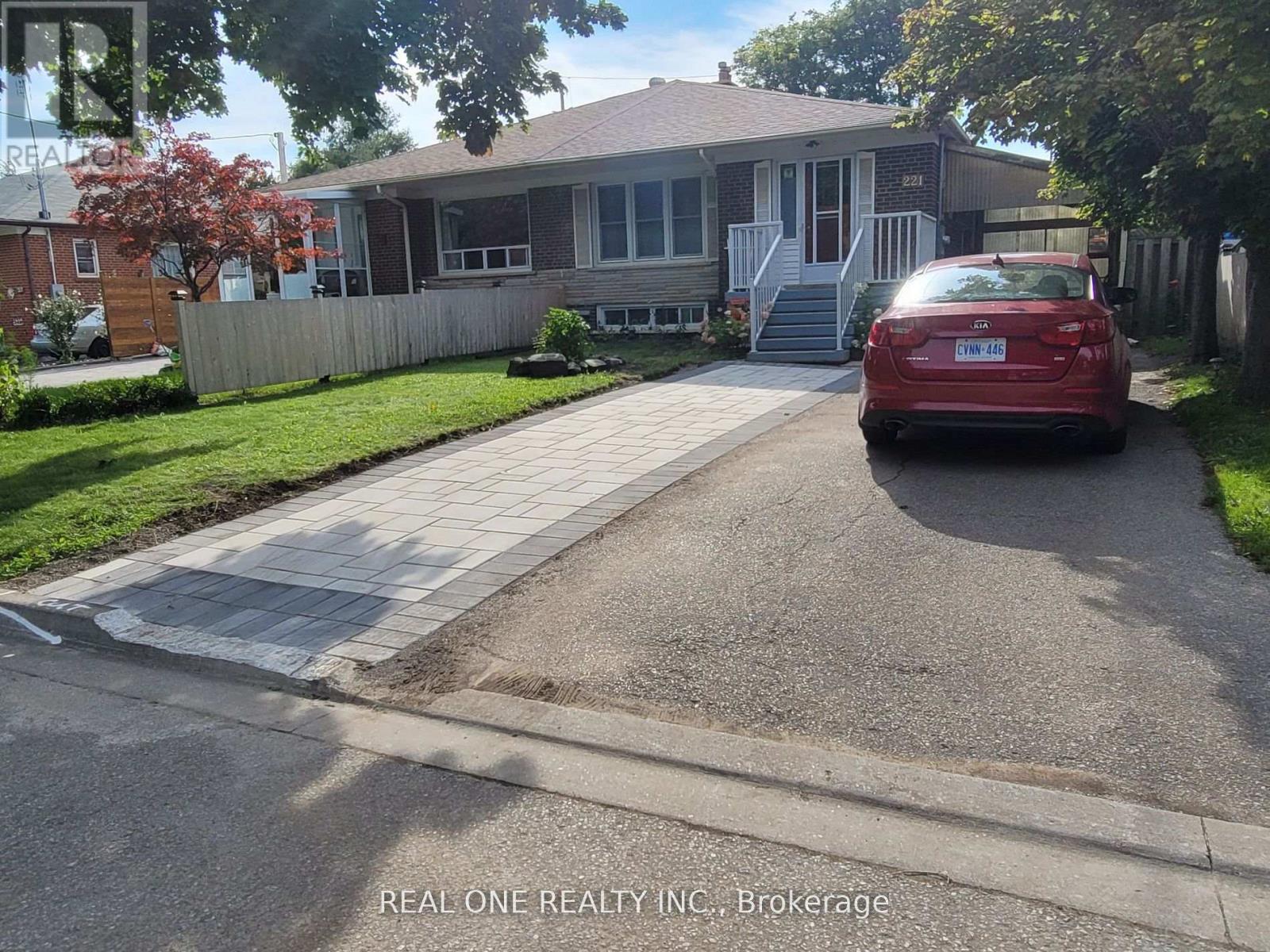 A red car is parked in front of a house with a sign that says "REAL ONE REALTY INC Brokerage.".