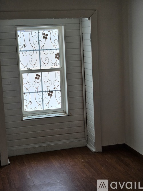 A window with a white frame and a decorative glass pane is in a room with wooden floors and white walls.