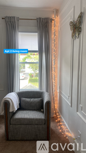 A grey armchair sits in a living room with a white wall and a window with grey curtains.